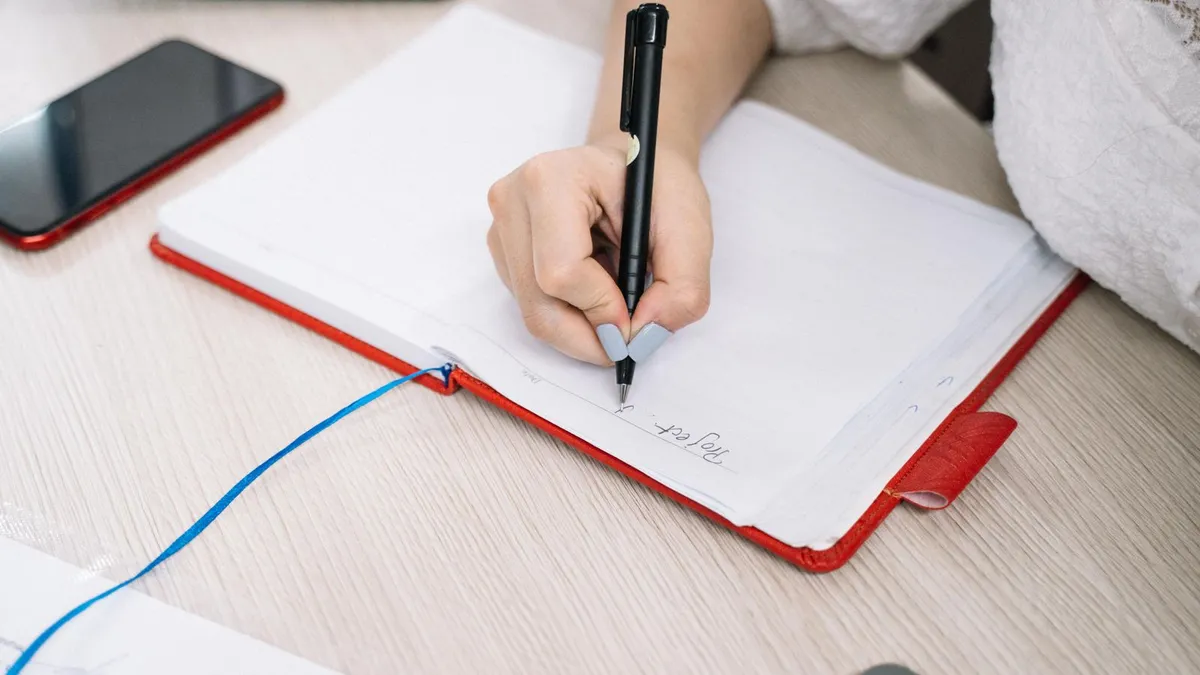 Person writing in a notebook beside a calculator and phone, keeping a register after they write a cheque